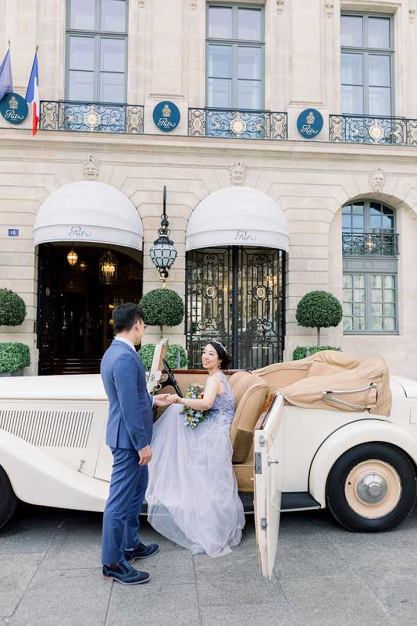 A couple poses in front of the Ritz Paris hotel on Place Vendôme, with the groom helping the bride step out of a cream-colored vintage convertible car. The bride wears a lavender-blue tulle ballgown with a lace bodice and holds a small bouquet featuring blue and mixed-color flowers, while the groom is dressed in a light blue suit. The shot is a full-length portrait taken at street level, capturing both figures and the iconic Ritz Paris facade with its signature teal logo medallions, white awnings, and French flag in the background. The overall styling has a classic Parisian aesthetic with the vintage vehicle serving as a key prop.