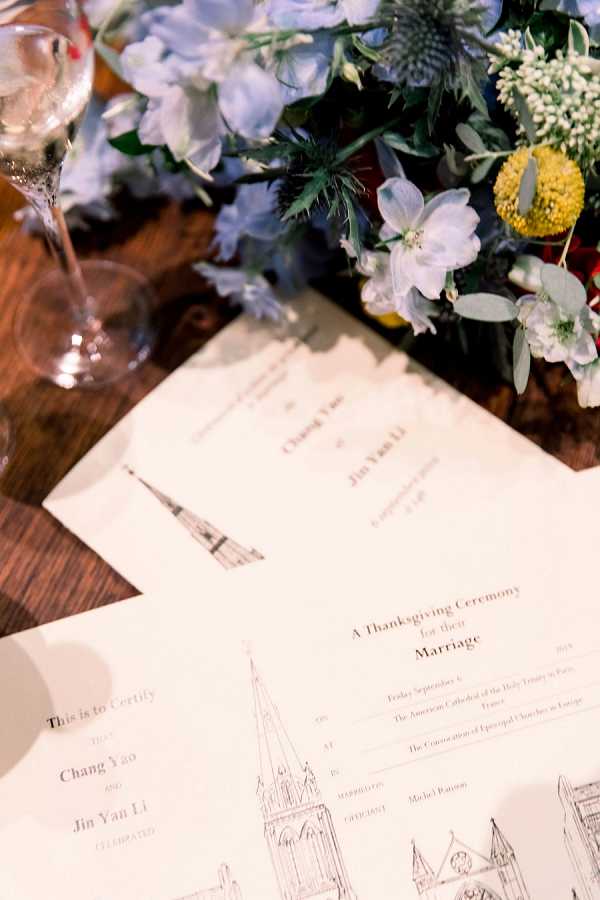 A close-up detail shot of wedding ceremony programs laid on a rustic wooden surface. The printed programs reference 'A Thanksgiving Ceremony for their Marriage' for Chang Yao and Jin Yan Li, held on Friday September 6 at the American Cathedral of the Holy Trinity in Paris, France, officiated by Michel Rannou. The programs feature an illustrated line drawing of a cathedral with a spire. Alongside the programs sits a floral arrangement featuring pale blue delphinium, blue thistle (eryngium), yellow craspedia (billy balls), white blooms, and eucalyptus, along with an empty stemmed wine glass, suggesting a reception table setting. The overall styling palette combines blue, yellow, and white florals with natural wood tones.
