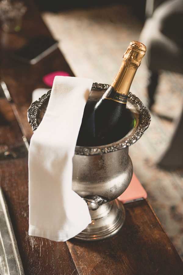 Close-up detail shot of a champagne bottle chilling in an ornate silver ice bucket with decorative embossed edging, placed on a dark wood surface. A white linen napkin is draped over the side of the bucket. The bottle has a gold foil top and a dark label. The background is softly blurred, showing what appears to be upholstered furniture and scattered items in warm, muted tones. The styling leans classic with the antique-finish silverware and white linen.