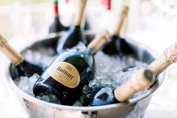 Close-up detail shot of multiple bottles of Ruinart Brut champagne chilling in a large silver ice bucket filled with crushed ice. The dark green glass bottles with gold labels are arranged horizontally and upright within the bucket, with additional bottles visible in the soft-focus background. The shot is taken at an outdoor or covered setting with bright natural light filtering in from behind. This appears to be a cocktail hour or reception beverage station setup.