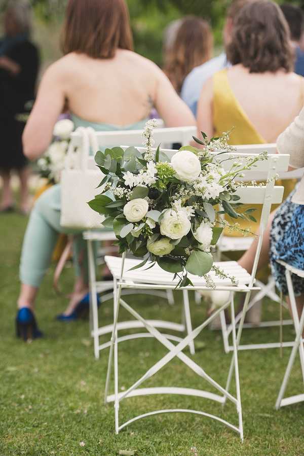 An outdoor wedding ceremony scene focused on a white metal folding bistro chair in the foreground, decorated with a floral arrangement of white ranunculus, small white blooms, and abundant green eucalyptus foliage tied to the chair back as an aisle marker. Several guests are seated in matching white bistro chairs in the background, dressed in casual summer attire including mint green trousers, a mustard yellow dress, and blue heeled shoes, suggesting a relaxed garden ceremony aesthetic. The decor palette is white and green with a garden-party style. Close-up portrait composition with a shallow depth of field that keeps the floral arrangement sharp while blurring the guests behind.
