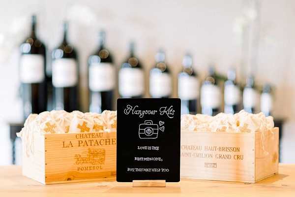 A close-up detail shot of a wedding reception favor display set on a wooden surface. Two wooden wine crates — one labeled 'Chateau La Patache, Pomerol' and the other 'Chateau Haut-Brisson, Saint-Emilion Grand Cru' — are filled with white tissue paper and used to hold hangover kit favors for guests. A small black chalkboard-style sign in the center reads 'Hangover Kits' with the phrase 'Love is the best medicine, but this may help too.' A row of dark wine bottles is softly blurred in the background. The styling is classic and wine-region-themed, incorporating authentic French Bordeaux wine crates as decor elements, consistent with a chateau or vineyard wedding setting.