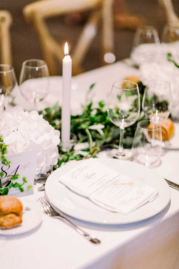 Close-up detail shot of a wedding reception table setting featuring a white linen tablecloth, stacked white porcelain plates topped with a printed menu card and a personalized place card reading 'Dorothy' in gold script. A lit white taper candle rises from a lush greenery runner composed of trailing foliage, accompanied by white hydrangea arrangements in white cube vases. A table number card (number 8) is visible on the left. Crystal wine glasses, silver cutlery, and bread rolls on side plates complete each place setting. The overall decor palette is white and green with gold accents, reflecting a classic, clean aesthetic.