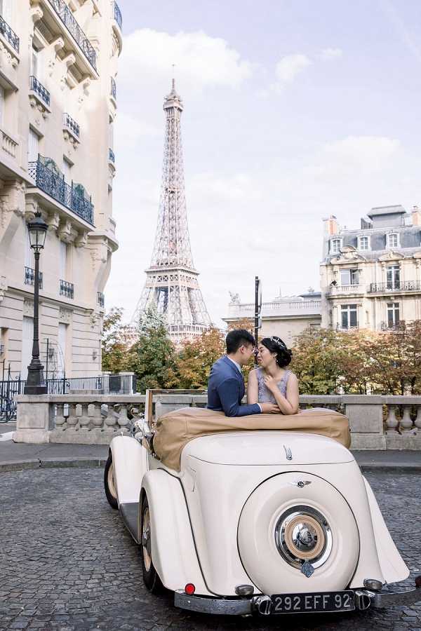 A couple poses in a portrait shot seated in the back of a cream-colored vintage convertible car on a cobblestone street in Paris, with the Eiffel Tower visible in the background between Haussmann-style buildings. The groom wears a navy blue suit and the bride wears a lavender lace dress with her hair up and a decorative headpiece; they are leaning toward each other as if about to kiss. The vintage car features tan leather upholstery and a rear-mounted spare wheel, adding a classic Parisian aesthetic to the shoot. The image is in color with warm, natural daytime lighting.