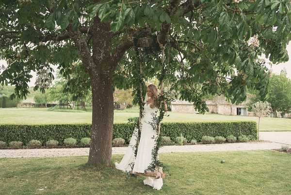 A bride sits alone on a wooden tree swing hanging from a large mature tree in the grounds of what appears to be a French country estate or chateau, visible in the background. The swing's ropes are wrapped with trailing greenery foliage, creating a decorative boho styling detail. The bride wears a fitted, long-trained white gown with a low back, and her hair is loosely down. The shot is a full-length outdoor portrait taken from a medium distance, capturing the bride looking downward with her dress and train pooling on the lawn around her.