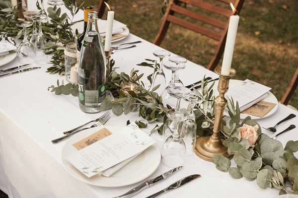 Close-up detail shot of an outdoor wedding reception table set with a white linen tablecloth. The centerpiece features a trailing greenery garland of eucalyptus and olive branches running the length of the table, accented with a single blush pink rose and small votive candles. Brass candlesticks holding white taper candles are placed along the garland, alongside clear glass water bottles and stemmed wine glasses turned upside down. Each place setting includes a white ceramic plate, silver cutlery, and a printed paper menu card with a kraft paper tag. Wooden folding chairs are visible in the background. The overall decor palette is white, gold, and greenery with a rustic-natural styling theme.