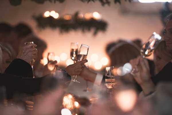 Guests raise champagne and wine glasses in a toast during an indoor wedding reception dinner. The setting is warmly lit with soft candlelight and warm-toned string lights or tea lights creating a bokeh-heavy background. A garland of dark greenery is suspended along the back wall, adding a decorative element to the space. The image is a mid-range shot with shallow depth of field, keeping the raised glasses in focus while guests' faces remain softly blurred; approximately 8–10 guests are partially visible around what appears to be a seated dinner table.