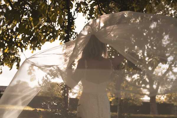 An outdoor bridal portrait showing a bride from behind as her long ivory veil billows and lifts dramatically in the wind, partially obscuring the frame. She is wearing a white or ivory gown with an open or low back, and her hair is worn down. The shot is taken in warm golden backlight, creating a soft, hazy, sun-flared exposure. The composition is a medium portrait taken from behind, with the sweeping veil filling much of the frame.