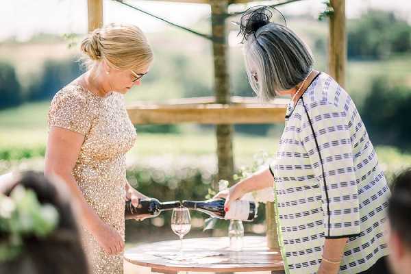 Two women are pouring wine at what appears to be an outdoor cocktail hour or reception, leaning over a small round wooden table with wine glasses on it. The woman on the left wears a gold sequined short-sleeve dress with her blonde hair up, while the woman on the right has grey and dark hair styled in an updo with a black fascinator and wears a white and blue striped jacket. The setting is an outdoor terrace or garden area with a wooden pergola structure visible in the background and green countryside beyond. The image is a mid-range portrait shot taken in bright natural sunlight.