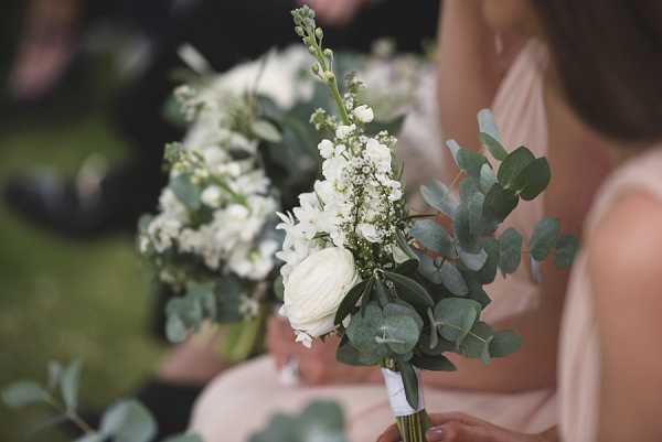 Close-up detail shot of a bridesmaid bouquet held during what appears to be an outdoor wedding ceremony. The bouquet features white ranunculus, white stock flowers, baby's breath, and eucalyptus foliage with silver-dollar eucalyptus leaves, wrapped with a white ribbon stem. A second similar bouquet is partially visible in the background. The bridesmaid holding the bouquet is wearing a blush pink dress. Seated guests are softly blurred in the background.