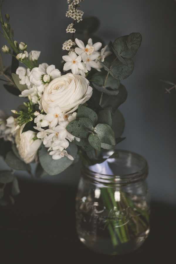Close-up detail shot of a floral arrangement displayed in a clear glass mason jar, set against a dark grey background. The arrangement features ivory ranunculus as the focal bloom, surrounded by small white star-shaped flowers, delicate white spray blooms, and silver-dollar eucalyptus foliage. The rustic-style presentation in a mason jar suggests a relaxed, organic aesthetic with a white and green color palette. The moody, low-key lighting adds strong contrast between the pale blooms and the dark background.