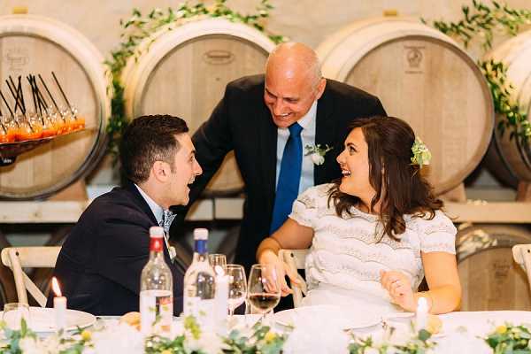 The bride and groom are seated at the head table during a reception, laughing and looking up at a man in a dark suit with a blue tie and boutonniere who is leaning in to speak with them. The setting is a winery barrel room, with large wooden wine barrels stacked along the back wall decorated with trailing green ivy garlands. The bride wears a white short-sleeved lace dress with a small floral hair accessory, while the groom wears a dark suit with a patterned tie. The table is set with white linens, wine glasses, water bottles, candles, and low greenery centerpieces with small yellow and white flowers. A food display with skewered appetizers is visible in the background to the left. The shot is a medium close-up portrait capturing a candid, joyful moment among the three.