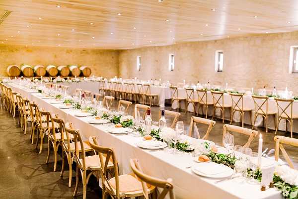 A wedding reception setup inside a winery chai or barrel room, with rows of oak wine barrels visible along the left wall. The space features warm stone walls, a wood-paneled ceiling with recessed lighting, and a polished concrete floor. Two long banquet tables are dressed in white linens and lined with natural wood cross-back chairs, set to accommodate a large number of guests. The table runners consist of lush greenery garlands with white blooms, complemented by white taper candles, white place settings, and wine glasses at each seat. The overall decor palette is white and green with a rustic yet clean aesthetic. Wide shot showing the full length of the reception space. Potential venue feature image.