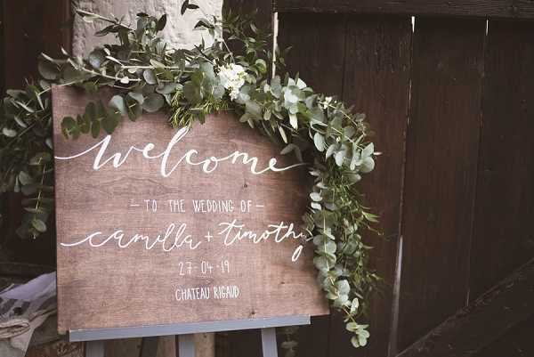 Close-up detail shot of a wooden welcome sign displayed on an easel, hand-lettered in white calligraphy script reading 'Welcome to the Wedding of Camilla + Timothy, 27-04-19, Chateau Rigaud.' The sign is decorated with a trailing garland of eucalyptus and small white flowers draped across the top and down one side. The styling is rustic with a natural, organic aesthetic, combining the raw wood panel with lush greenery. The sign is positioned against a dark wooden backdrop.