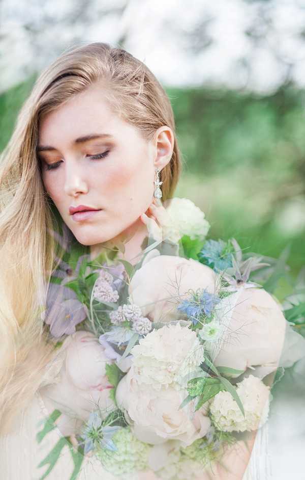 A close-up bridal portrait of a woman with long blonde hair, eyes downcast, wearing a crystal drop earring and natural makeup with a warm blush tone. The image uses a double-exposure technique, layering the bride's face with a bouquet of blush peonies, ivory hydrangeas, pale blue nigella, soft purple clematis, and green foliage, creating a soft, painterly overlap of the two images. The overall color palette is muted and pastel — blush, ivory, sage, and powder blue. The composition is a tight portrait shot with a shallow depth of field and a light, airy feel.