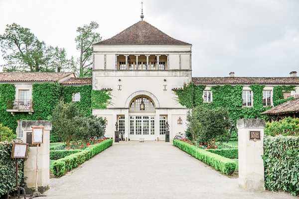 Wide exterior shot of a French château venue featuring a symmetrical façade with ivy-covered wings flanking a central white rendered tower with a steeply pitched hip roof. The main entrance is framed by a large arched opening with white French doors and a lantern above, approached by a straight gravel driveway lined with neatly trimmed low box hedging and small planted borders. No people are visible in the frame. The image has a soft, slightly overcast light giving it a muted, pale tone. Potential venue feature image.