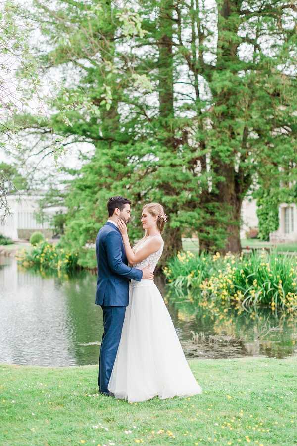 A couple portrait taken outdoors on the grounds of what appears to be a French château or manor, positioned beside a calm ornamental pond. The bride wears a white tulle A-line gown with a lace-detailed bodice and has her hair pinned up with a small floral hair accessory, while the groom wears a navy blue suit. The two stand facing each other in an intimate pose, with the bride's hand resting on the groom's jaw. The stone façade of a building is partially visible through the trees in the background. The shot is a full-length portrait with soft, even natural lighting and a slightly overcast look, giving the image a light, airy feel.
