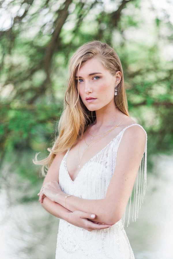 A close-up portrait of a bride standing outdoors with a softly blurred green tree and water background. She wears a white sleeveless wedding dress with a deep V-neckline featuring beaded fringe detail along the straps and bodice. Her accessories include layered gold necklaces, a crystal drop earring, and a delicate gold bracelet. Her blonde hair is worn loosely down with a subtle half-back pin. The composition is a tight mid-shot with shallow depth of field, and the overall styling leans toward a bohemian aesthetic.