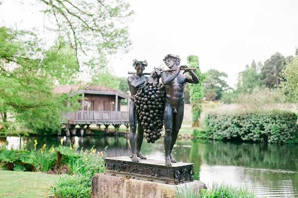 A close-up shot of a bronze sculpture depicting two classical figures carrying a large bunch of grapes on a pole between them, positioned on an ornate stone pedestal. The sculpture is set within formal garden grounds featuring a decorative wooden pavilion or boathouse structure over a calm pond in the background. No people or wedding party are visible in this image; the shot focuses entirely on the venue's decorative garden feature. Potential venue feature image.
