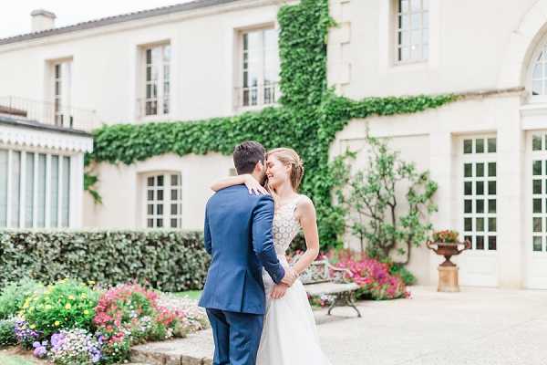 A couple portrait taken outdoors in the courtyard of a French chateau or manor house, with the two-story cream-rendered building prominently visible in the background covered in climbing ivy. The groom wears a navy blue suit and has his back partially to the camera, while the bride faces him smiling, dressed in a white ballgown with an embellished lace bodice and her hair pinned up. They are holding hands and the bride has her arm around the groom's shoulder in an intimate pose. The composition is a medium portrait shot with soft, even natural light and a slightly blurred background that keeps focus on the couple. Potential venue feature image.