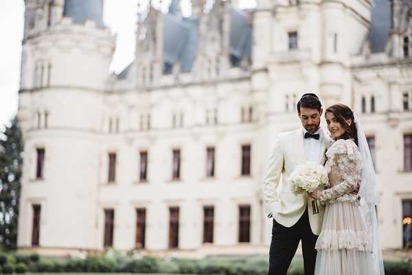 A couple poses together outdoors in front of a large French chateau with ornate Renaissance-style architecture, turrets, and slate rooftops. The groom wears a white dinner jacket with black trousers and a black bow tie, while the bride wears a long-sleeved lace and floral-embroidered gown with a long veil, holding a round bouquet of white roses and white blooms. The two are standing close together, the groom looking down toward the bride in a candid, relaxed portrait. The composition is a medium wide shot with the chateau facade centered and softly blurred in the background. Potential venue feature image.