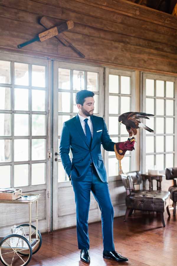 A groom stands alone indoors holding a harris hawk or similar bird of prey on a burgundy falconry glove on his right hand, looking down at the bird. He is wearing a fitted navy blue suit with a matching navy tie, white dress shirt, white pocket square, and black Oxford shoes. The setting is a rustic interior space with weathered white-painted French doors, exposed wooden ceiling beams, wide-plank hardwood floors, and vintage furnishings including a brass bar cart on the left and leather seating in the background. The portrait is a full-length shot with a classic, editorial feel, and the falconry element serves as a distinctive styled detail.