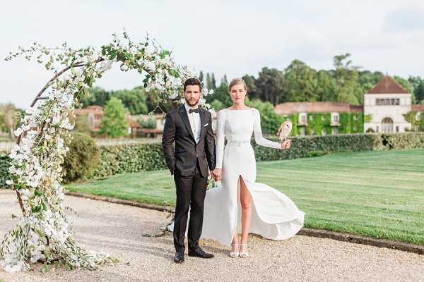 A couple poses outdoors on a gravel path in front of a circular floral arch decorated with white blooms and trailing greenery. The groom wears a black tuxedo with a bow tie, while the bride wears a sleek long-sleeve white gown with a front slit, a embellished belt at the waist, and holds a barn owl on her gloved arm. The setting appears to be the manicured grounds of a French chateau or manor house, visible in the background with ivy-covered walls and formal lawns. The shot is a full-length portrait with a classic, editorial style.