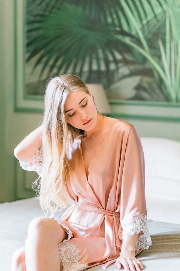 A getting-ready portrait of a bride sitting on a white bed, wearing a blush satin robe with white lace trim at the sleeves and hem. She has long blonde hair worn loosely down and is looking downward with one hand raised to her hair. The indoor setting features a botanical palm-print mural or wallpaper in deep green tones visible behind her, along with a lamp on a bedside table. The shot is a close-up portrait with soft, bright natural light and a clean, romantic getting-ready aesthetic.