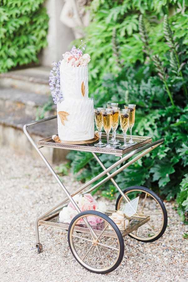 A styled detail shot of a gold vintage bar cart displayed outdoors on a gravel path, set against lush garden greenery with a stone statue partially visible in the background. The upper shelf holds a two-tier white textured wedding cake decorated with gold leaf accents, lavender and blush florals including what appear to be macarons and small blooms on top, alongside five filled champagne flutes. The lower shelf contains a loose arrangement of blush and dusty pink peonies along with a small triangular paper decoration. The overall styling is classic-French with gold and blush tones, and the image is a medium wide shot taken in bright natural daylight.