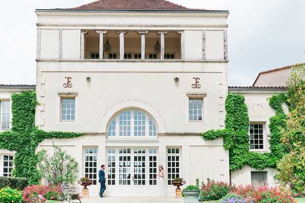 A couple poses in front of a large white neoclassical French chateau facade, partially covered in climbing ivy, with tall French doors and arched windows on the ground floor and a colonnaded upper balcony. The bride wears a white gown and the groom wears a dark navy suit; they stand facing each other at a slight distance on the terrace in what appears to be a candid walking or first-look moment. Ornamental terracotta urns and colorful flower beds flank the entrance. Wide shot composition emphasizing the scale of the building relative to the couple. Potential venue feature image.