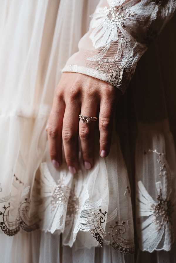 A close-up detail shot of a bride's hand resting against her wedding dress, showcasing a rose gold engagement ring with a round center stone and a matching diamond-set band. The bride's nails are painted a pale blush pink. The dress features intricate ivory lace with embroidered floral and scrollwork motifs, crystal or bead embellishments, and long sheer sleeves with scalloped lace cuffs. The layered skirt of the dress is visible in the lower portion of the frame, with matching lace appliqué detailing along the hem. The overall styling is romantic and vintage-inspired.