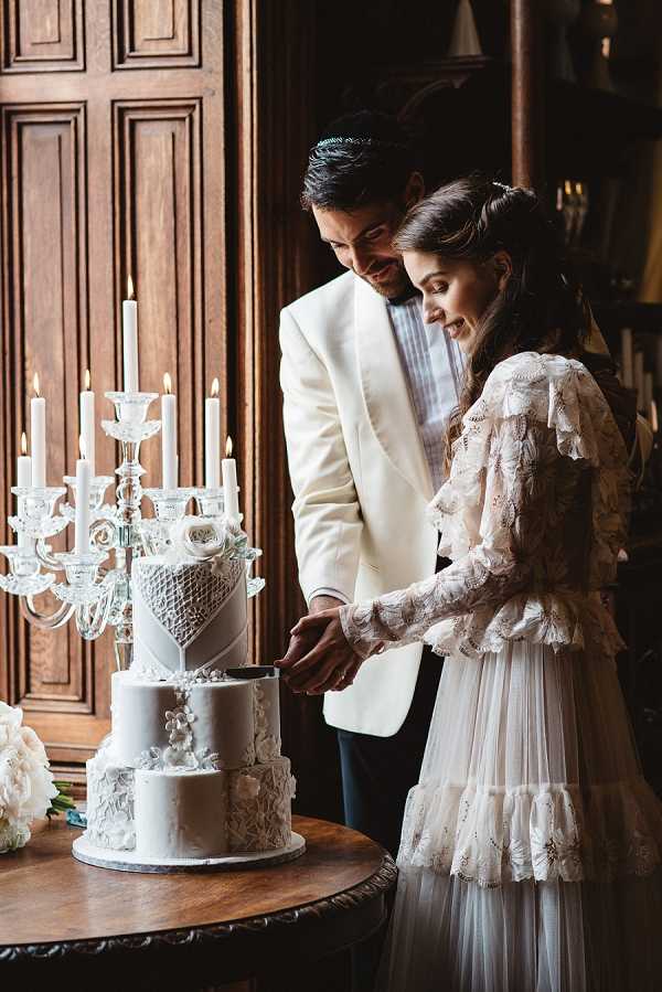The bride and groom are cutting their wedding cake indoors, set against a backdrop of dark wood paneling that suggests a classic interior, likely a chateau or manor library. The groom wears a white dinner jacket with dark trousers and a kippa, while the bride wears a long-sleeved lace and tulle dress with ruffle-tiered detailing in ivory. The three-tier wedding cake is all-white with textured fondant decorations including floral appliqués, a lattice panel, and ribbon-style detailing, topped with a white rose. A crystal candelabra with lit white tapered candles stands beside the cake, and a white floral arrangement — appearing to include peonies — is partially visible to the left. The shot is a medium portrait framing both figures from approximately waist height, capturing their expressions as they smile downward toward the cake.