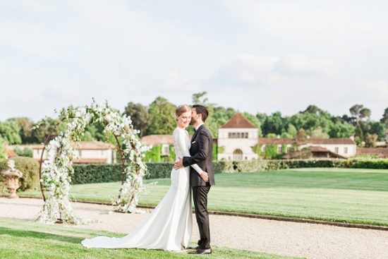 A bride and groom share an intimate moment outdoors on a formal garden lawn, with the groom leaning in to kiss the bride on the forehead or cheek. The bride wears a minimalist long-sleeve white gown with a fitted silhouette and a trailing train, while the groom is dressed in a dark charcoal suit. Behind them stands a circular floral arch adorned with white blooms and small green foliage, positioned along a manicured hedgerow pathway. A French-style estate building with a terracotta roof is visible in the background. The image is a medium-wide portrait shot with soft, airy natural light. Potential venue feature image.