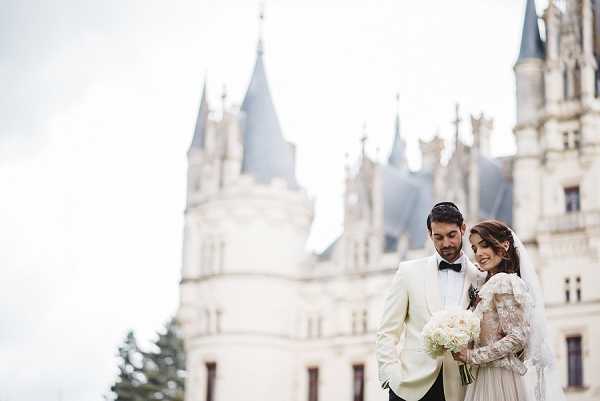 A couple portrait taken outdoors in front of a large French chateau with Gothic-style turrets and pale stone architecture visible in the soft-focus background. The groom wears a cream/ivory dinner jacket with a black bow tie and black trousers, and the bride wears a long-sleeved lace wedding gown with a veil, holding a compact bouquet of white flowers including what appear to be roses or peonies. The two are standing close together, both looking downward with relaxed expressions, in a candid-style portrait composition shot at mid-length. Potential venue feature image.