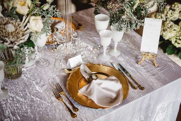 Close-up detail shot of a styled wedding reception place setting on a silver crushed velvet tablecloth. The place setting features a gold charger plate with a white folded napkin secured by a decorative cameo napkin ring, flanked by gold-handled cutlery including a fork, knife, and additional utensils. Two white milk glass goblets are positioned above the plate alongside crystal glassware and ornate candle holders. A small white place card and a gold-footed menu card holder are visible to the right. The floral centerpiece includes cream and ivory blooms — likely peonies and proteas — with dusty miller and eucalyptus foliage. The overall decor palette is gold, ivory, and silver with a classic, ornate styling theme.