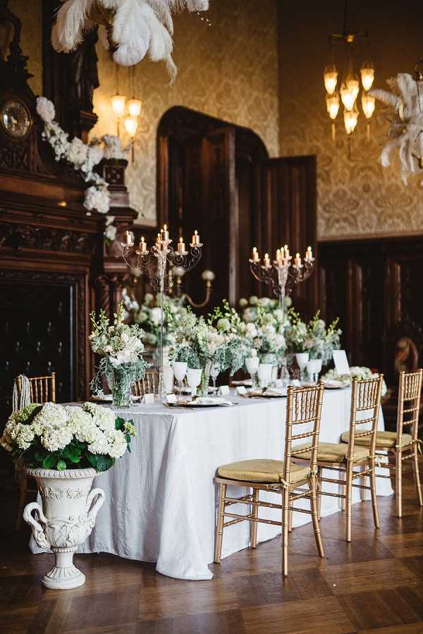 A reception tablescape detail shot taken inside a grand historic ballroom with dark carved wood paneling, patterned wallpaper, and warm chandelier lighting. The long rectangular dining table is dressed in a white linen cloth and set with gold chiavari chairs with gold cushions, white place settings, and gold-rimmed glassware. Centerpieces consist of low arrangements of white hydrangeas, cream roses, and soft greenery, accented by tall silver candelabras with lit ivory candles. A large white ornamental urn on the floor in the foreground holds a full arrangement of white hydrangeas and foliage. Overhead, white ostrich feather plumes hang as a decorative installation, and a large floral garland of white blooms drapes across the carved stone mantelpiece in the background. The overall decor palette is white, cream, and soft green with gold accents, reflecting a classic, formal styling theme. Potential venue feature image.