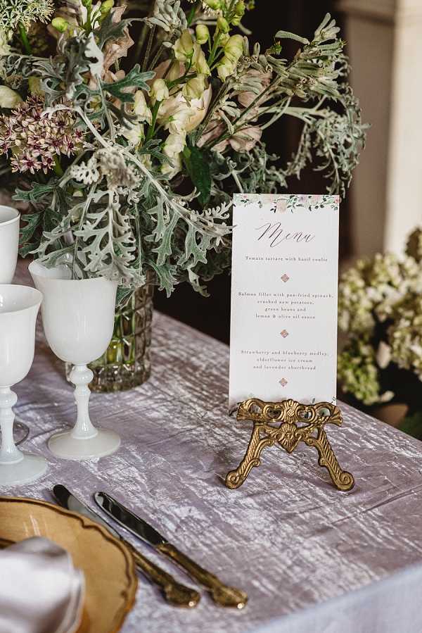 Close-up detail shot of a wedding reception table setting featuring a printed menu card displayed on an ornate gold easel stand. The menu card has floral watercolor accents at the top and lists courses including tomato tartare, salmon fillet with pan-fried spinach, and a strawberry and blueberry dessert. The table is dressed with a dusty lavender crushed velvet linen, and place settings include gold-rimmed charger plates, gold flatware, and white milk glass goblets. A lush floral arrangement in a glass vase sits behind, composed of blush snapdragons, dusty miller, cream blooms, and trailing greenery in a muted, romantic palette. The overall styling is vintage-romantic with a soft grey, blush, and gold color scheme.