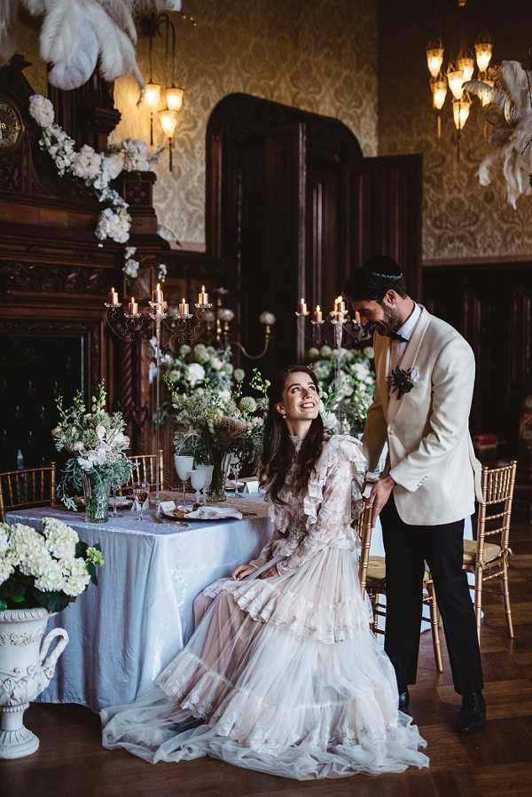A couple poses at a sweetheart table inside a grand ballroom or manor hall featuring dark wood-paneled walls, ornate wallpaper, and a large carved fireplace mantel. The bride is seated and wearing a blush-pink ruffled long-sleeve gown with tiered tulle layers, looking up at the groom who stands beside her wearing a cream/ivory dinner jacket with black trousers, a bow tie, and a boutonniere with dark foliage. The table is dressed in a pale blue-grey linen and styled with gold candelabras holding lit taper candles, low floral arrangements of white hydrangeas, ivory blooms, and mixed greenery, gold chiavari chairs, and crystal glassware. Large white feather plumes hang decoratively above, and a white ceramic urn filled with white hydrangeas sits on the floor in the foreground. The overall styling theme is vintage-romantic with a black, ivory, and blush palette, warm candlelight, and ornate period decor. Medium-wide portrait shot.
