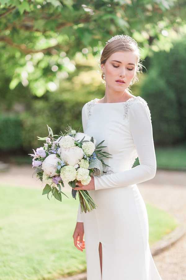 A bridal portrait taken outdoors in a garden setting, with a softly blurred green foliage background. The bride wears a long-sleeve ivory fitted gown with a bateau neckline, a front slit, and crystal embellishment detail at the shoulders, paired with a delicate crystal hair piece and drop earrings. She holds a medium-sized bouquet composed of white peonies, cream hydrangeas, lavender roses, blue thistle, and trailing greenery. The image is a close-up portrait shot with the bride glancing downward toward her bouquet.
