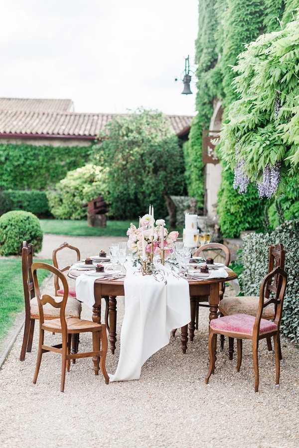 An outdoor tablescape styled for a wedding reception, set on a gravel courtyard in front of an ivy-covered stone building with a terracotta-tiled roof. The round dark wood dining table is dressed with a white linen runner and surrounded by six mismatched antique French wooden chairs, including rush-seat and dusty pink velvet-upholstered styles. The centerpiece features a brass candelabra with taper candles, flanked by an arrangement of blush and cream ranunculus with trailing greenery, and a tall white pillar candle beside champagne flutes. Place settings include dark charger plates, glassware, and small dark dessert items at each seat. The overall styling is romantic and rustic-classic, mixing vintage French furniture with a soft blush and gold color palette. Wide shot taken from a slight elevation angle showing the full table setting within its courtyard environment.