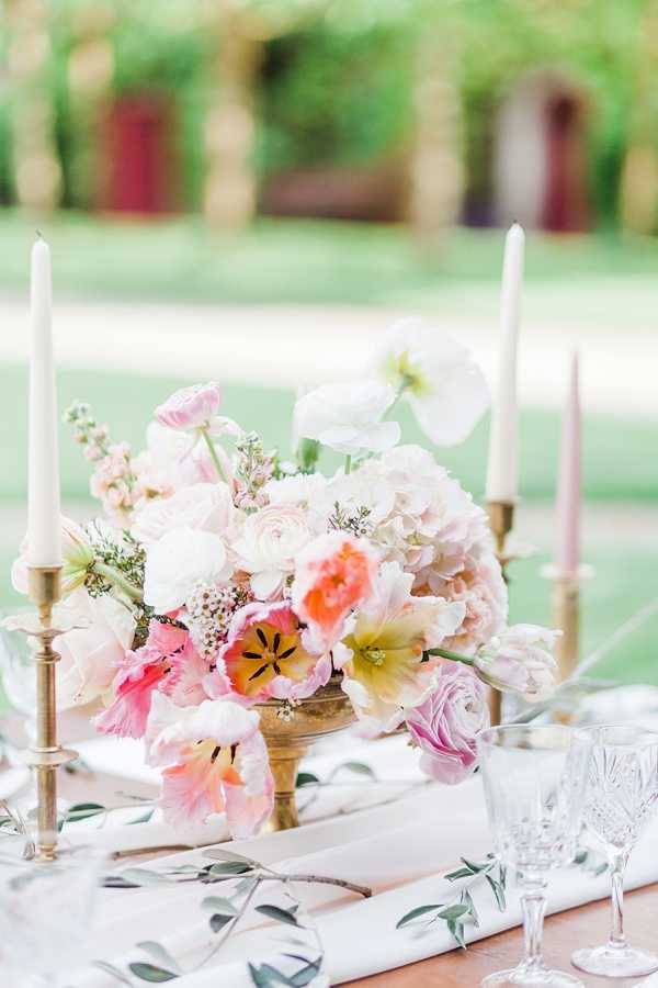 Close-up detail shot of an outdoor wedding reception table centerpiece. The arrangement is set in a brass pedestal compote vase and features a mix of blush pink ranunculus, white poppies, coral and pink parrot tulips, light pink hydrangea, and small wax flower accents in a soft, garden-style composition. Brass taper candlestick holders with white and blush pink unlit taper candles flank the centerpiece on both sides. The table is styled with white linen napkins printed with an olive branch motif and crystal cut-glass stemware. The overall decor palette combines brass, blush, coral, and white in a romantic garden aesthetic.