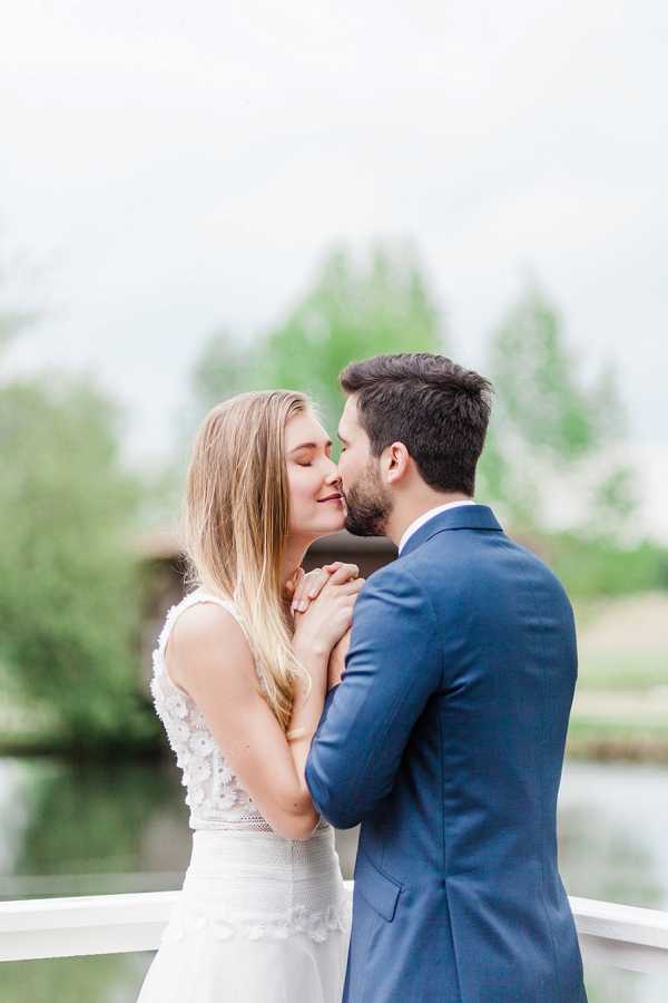 A couple portrait shot outdoors, with the bride and groom sharing a close moment, nearly kissing, while holding hands between them. The bride wears a sleeveless white gown with 3D floral lace appliqué detailing on the bodice and long blonde hair worn down, while the groom wears a navy blue suit with a light blue dress shirt. They are standing near a white railing with a softly blurred water feature and trees visible in the background. The composition is a medium portrait shot with a shallow depth of field, giving the image a light, airy feel.