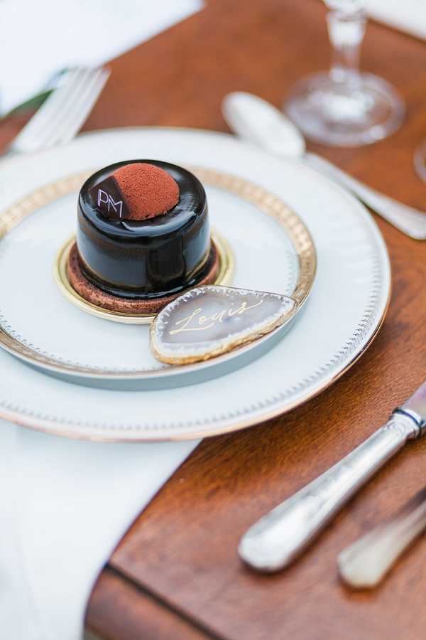 Close-up detail shot of a wedding reception place setting on a warm-toned wooden table. A white dinner plate with a gold beaded rim sits on a matching gold-rimmed charger, holding a small individual chocolate dessert with a dark mirror-glaze finish and a cocoa-dusted dome topping, monogrammed with 'PM' in white. A grey agate slice place card with the name 'Louis' written in gold calligraphy is propped against the dessert. Silver cutlery and a crystal wine glass are visible in the background, suggesting a classic, refined reception table design with a dark chocolate, gold, and grey decor palette.