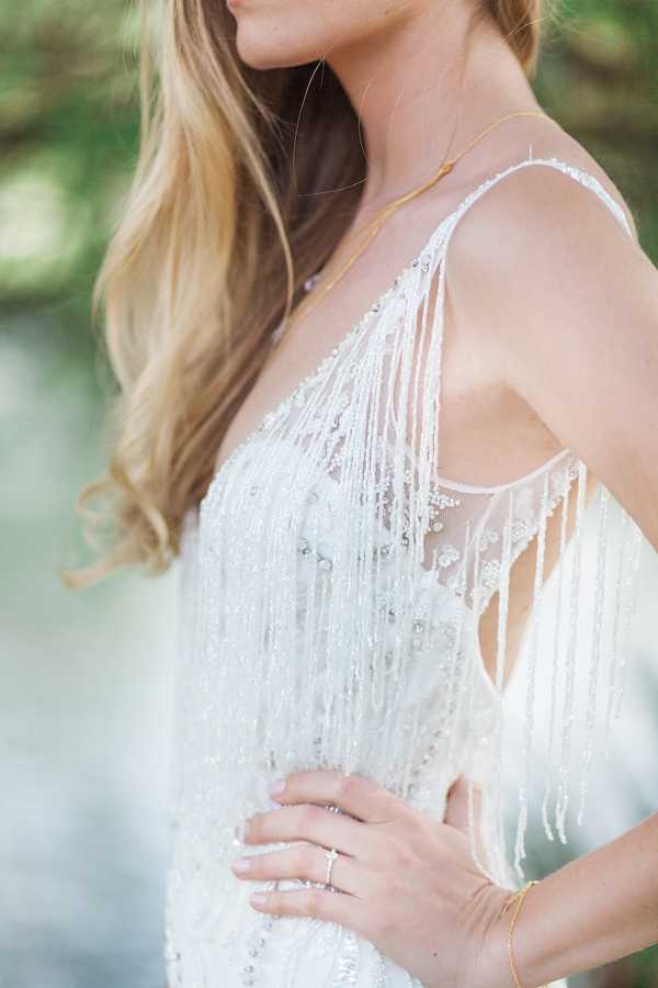 Close-up portrait of a bride shot outdoors with a softly blurred natural background. The image focuses on the bodice and neckline of a white beaded and sequined wedding dress featuring heavily embellished fringe detailing that falls vertically across the front and over the shoulders, creating a 1920s-inspired Art Deco aesthetic. The bride has long, loosely waved blonde hair and wears a delicate gold chain necklace, a thin gold bracelet, and a diamond ring. Her hand rests on her hip, partially showing the ring and bracelet details.