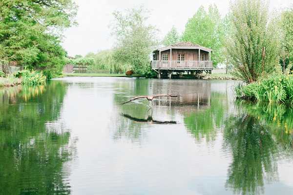 Wide-shot image of a wooden pavilion or gazebo structure built on stilts over the edge of a calm pond, surrounded by lush green trees and waterside vegetation. A large bird — appearing to be a heron — is captured mid-flight just above the water's surface in the foreground, its wings spread wide and touching the water. The pavilion features a wraparound deck and a tiled roof, and its reflection is visible in the still water below. No people or wedding-specific decor are visible in the frame. Potential venue feature image.