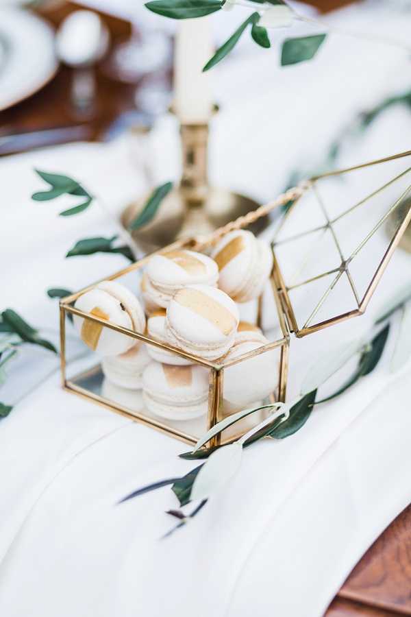 Close-up detail shot of a wedding reception tablescape featuring ivory and gold-brushed French macarons displayed inside an open geometric brass and glass terrarium box. The table is dressed with a white linen runner and scattered greenery sprigs, with a brass taper candle holder and white candle visible in the soft-focus background. The overall decor palette is white, gold, and green, consistent with a modern minimalist styling approach. A second geometric brass terrarium is partially visible to the right.