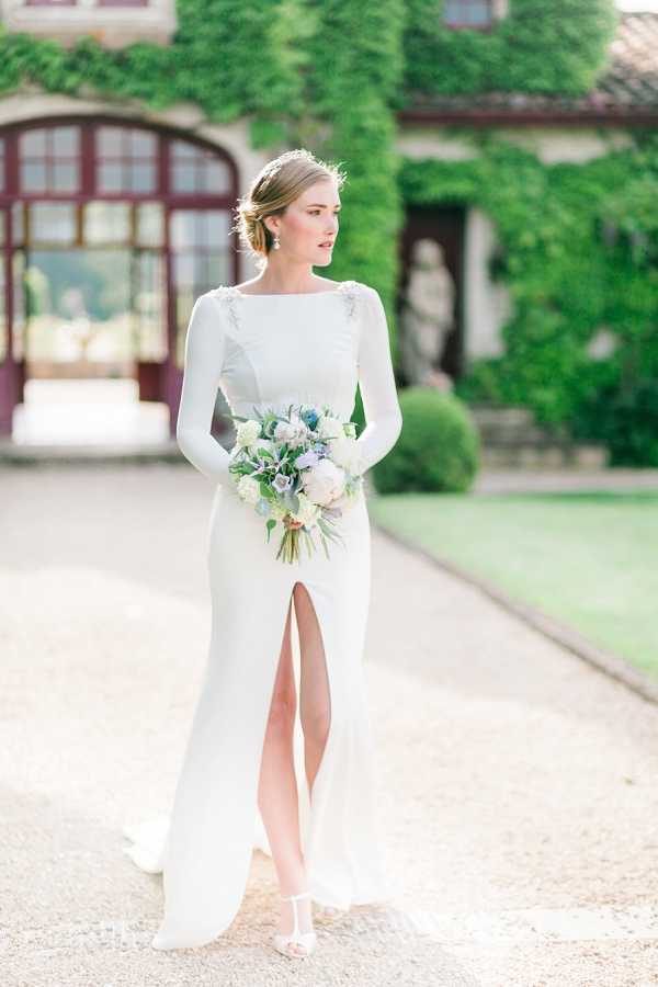A bridal portrait taken outdoors on a gravel driveway in front of a ivy-covered manor or chateau facade with arched dark-wood windows. The bride stands alone, gazing to one side, wearing a long-sleeve, fitted ivory crepe gown with a high bateau neckline, a front slit, and small embellished details at the shoulders. She holds a medium-sized bouquet composed of white garden roses, pale blush blooms, dusty blue thistles, soft lavender flowers, and trailing eucalyptus foliage. Her hair is pulled back into a low updo with a delicate headpiece, and she wears drop earrings. The styling is clean and modern-classic. The shot is a full-length portrait with soft, even natural light and a shallow depth of field that blurs the background architecture.