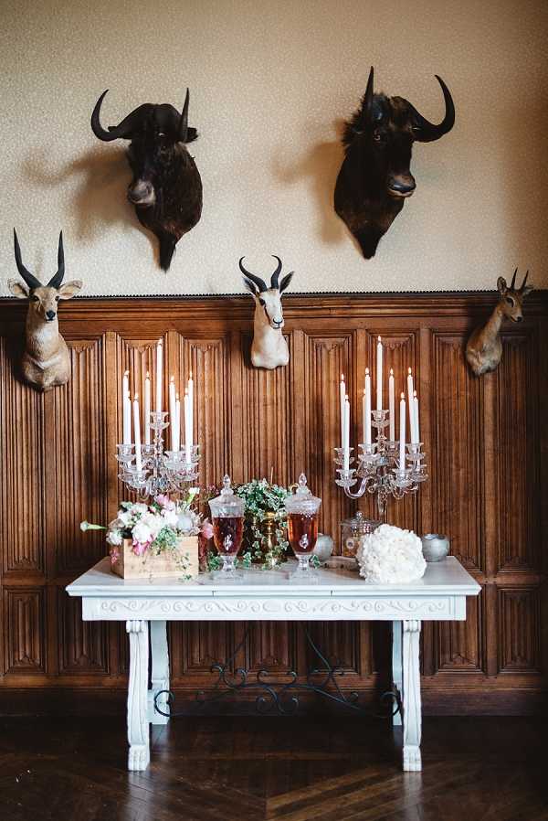 A styled wedding reception display table photographed indoors against a dark wood-panelled wall featuring five taxidermy animal mounts, including two large buffalo and three antelope or gazelle heads. The white ornate table holds two tall crystal candelabras with white taper candles, glass apothecary jars filled with amber liquid, trailing ivy, a wooden crate with blush pink and white floral arrangements including what appear to be peonies and roses, and a white hydrangea or carnation floral arrangement on the right. The overall decor palette mixes rustic and classic French chateau styling. Wide, symmetrically composed shot showcasing the full table vignette. Potential venue feature image.