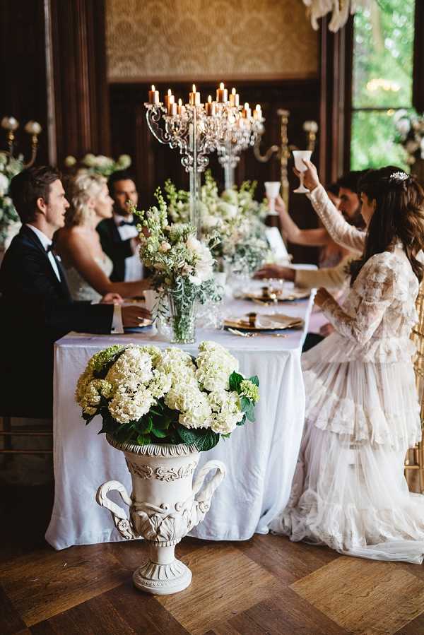 A toast is being made at an indoor wedding reception dinner, set in a wood-paneled room with ornate gold-toned wall detailing suggesting a historic chateau or manor. Four guests are visible seated at a rectangular table covered in a pale blue-grey linen, set with gold charger plates, while a woman in a tiered ivory lace long-sleeve gown raises a glass in the foreground. The table is decorated with tall crystal candelabras holding lit ivory taper candles, alongside low arrangements of white and blush flowers with greenery. In the foreground, a large ornate white urn vase holds a full arrangement of cream hydrangeas and foliage on the parquet wood floor. The overall decor palette is white, cream, and gold with a classic, formal styling. Medium-wide shot taken from a low angle.