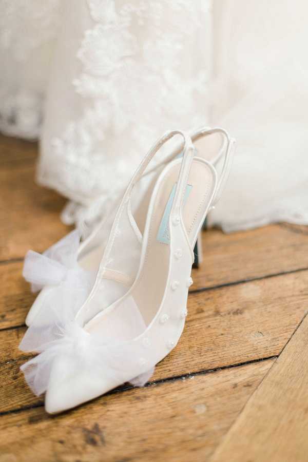 Detail close-up shot of a pair of white bridal slingback heels placed on a rustic wooden floor, with the hem of a lace wedding dress visible in the soft-focus background. The shoes feature sheer mesh panels, small pearl embellishments along the straps, tulle bow details at the pointed toe, and a pale blue insole — a nod to the 'something blue' tradition. The composition is a styled flat-lay-style detail shot with warm, soft natural lighting.
